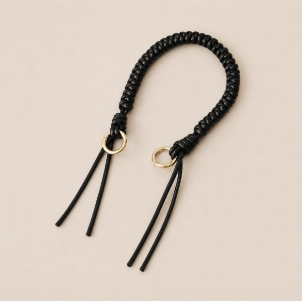 Folie à Trois Layover short wholesale phone strap in black braided cord with gold rings, shown as a knotted loop with fringe ends on a cream background.