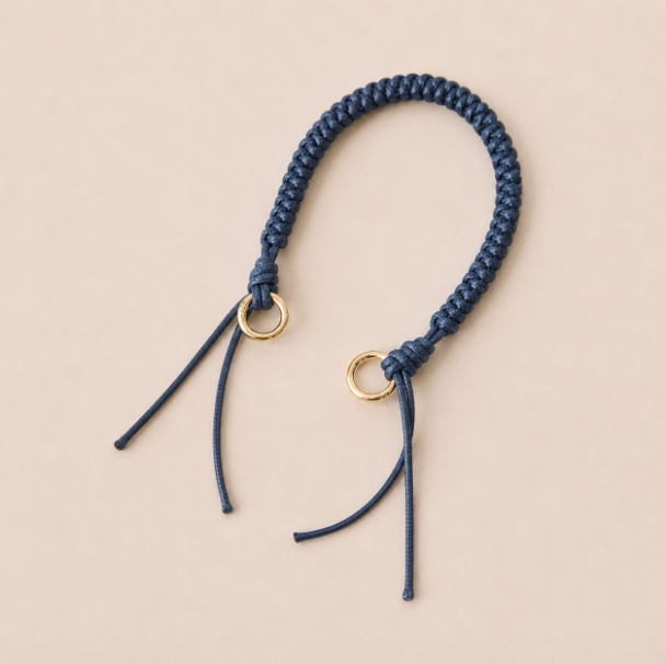 Folie à Trois Layover short wholesale phone strap in navy blue braided cord with gold rings, shown as a knotted loop with fringe ends on a cream background.