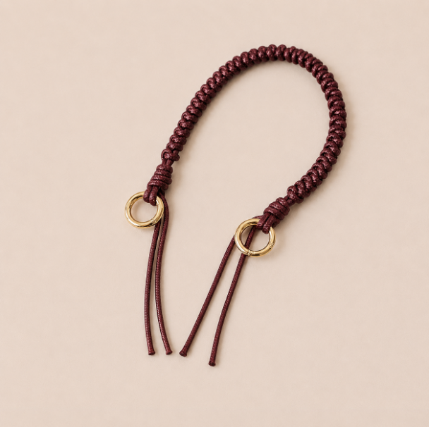 Folie à Trois Layover short wholesale phone strap in burgundy braided cord with gold rings, shown as a knotted loop with fringe ends on a cream background.