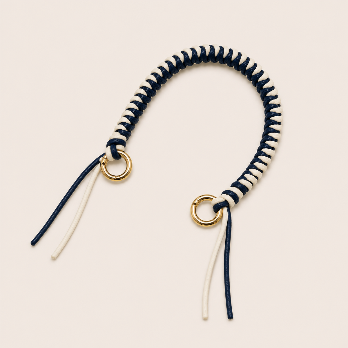 Folie à Trois Layover short wholesale phone strap in navy and white braided cord with gold rings, shown as a knotted loop with fringe ends on a cream background.