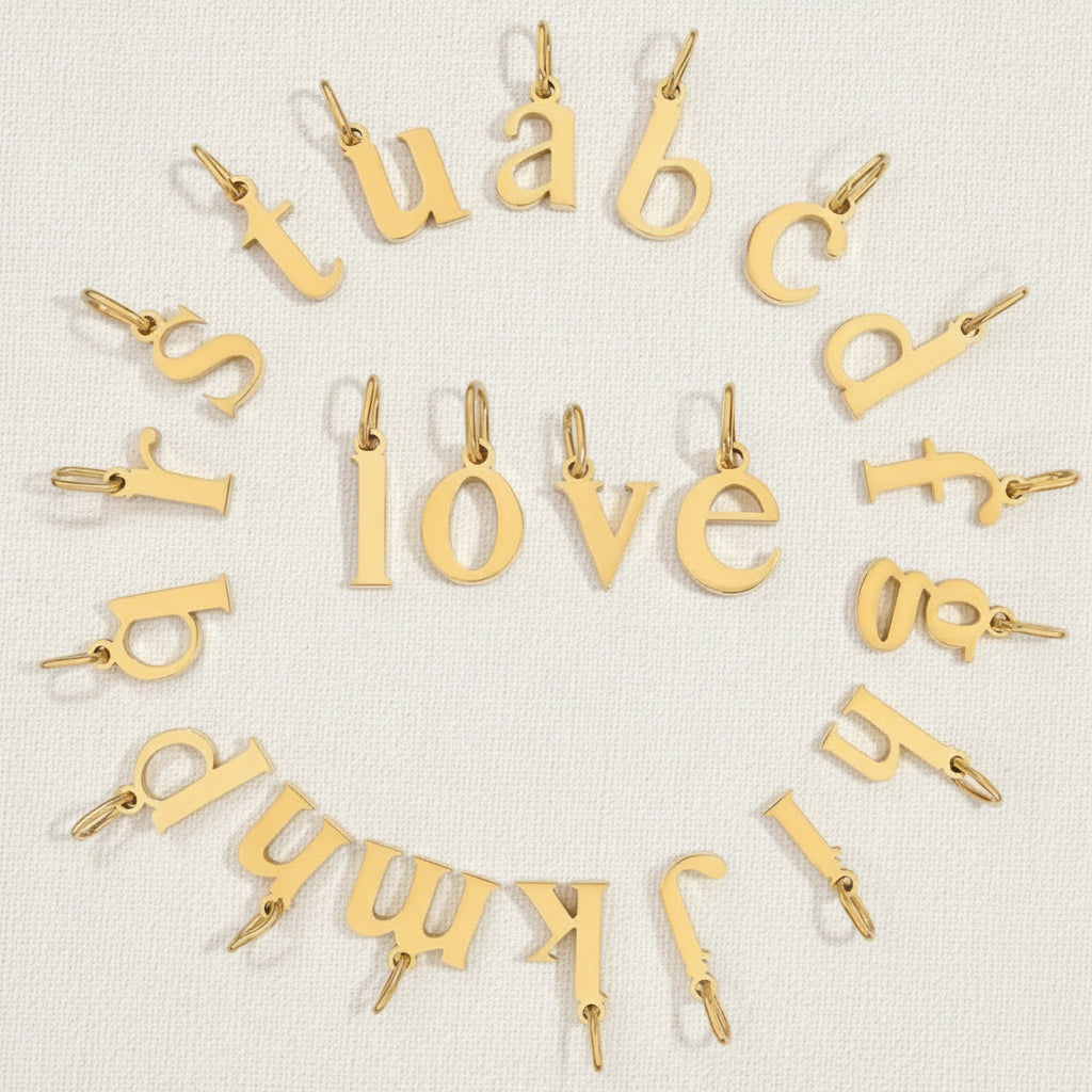 The Letter CHARM collection in a circular flat lay spelling 'love' with scattered gold lowercase letters on tan textured fabric.