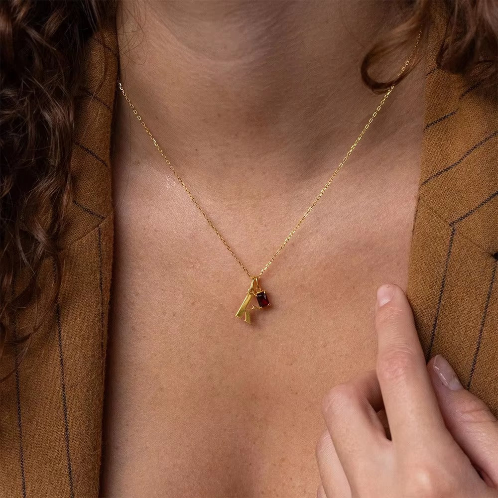 The Letter CHARM worn at the neckline, a single gold lowercase letter charm set with a small red gem, layered on a pinstripe blazer.