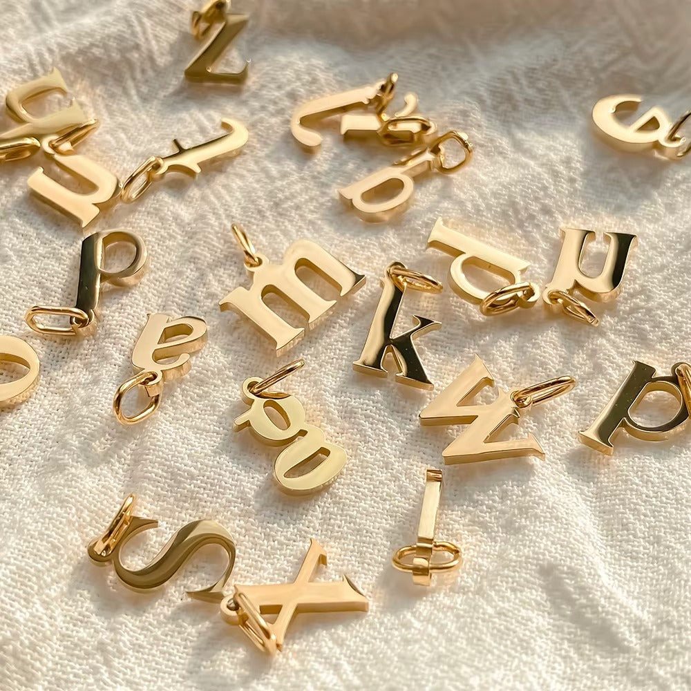 The Letter CHARM collection in close-up, dozens of gold lowercase letter charms scattered together on white textured fabric.