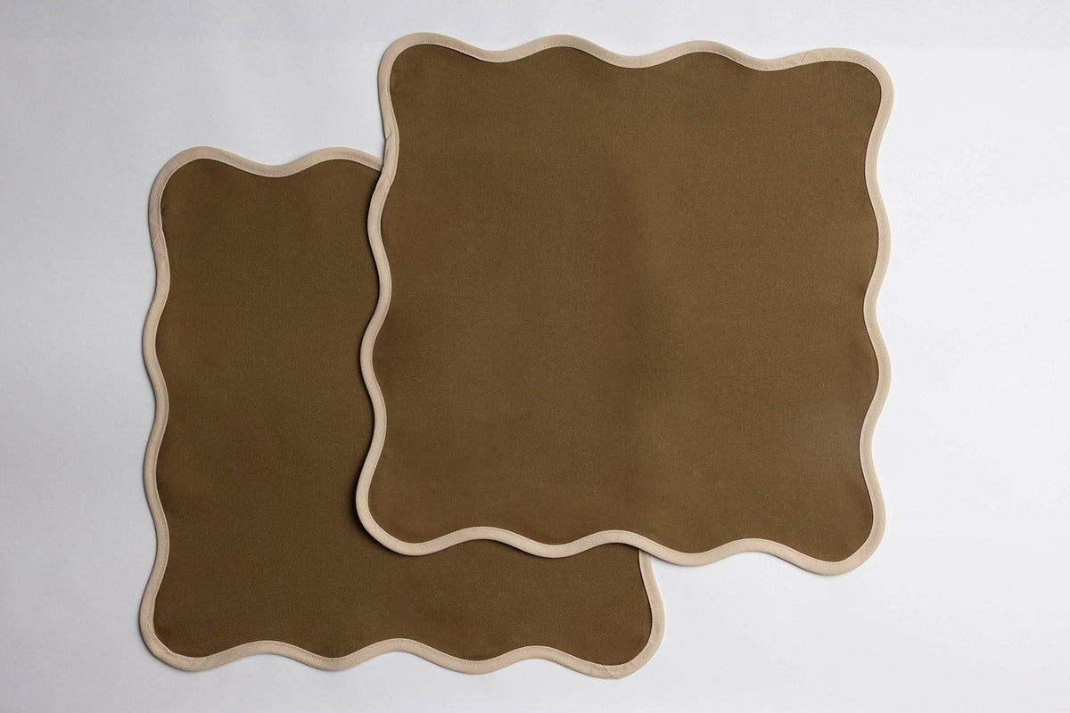 Two olive green Lilou Scallop napkins edged with a cream scalloped border, laid flat and overlapping on white.