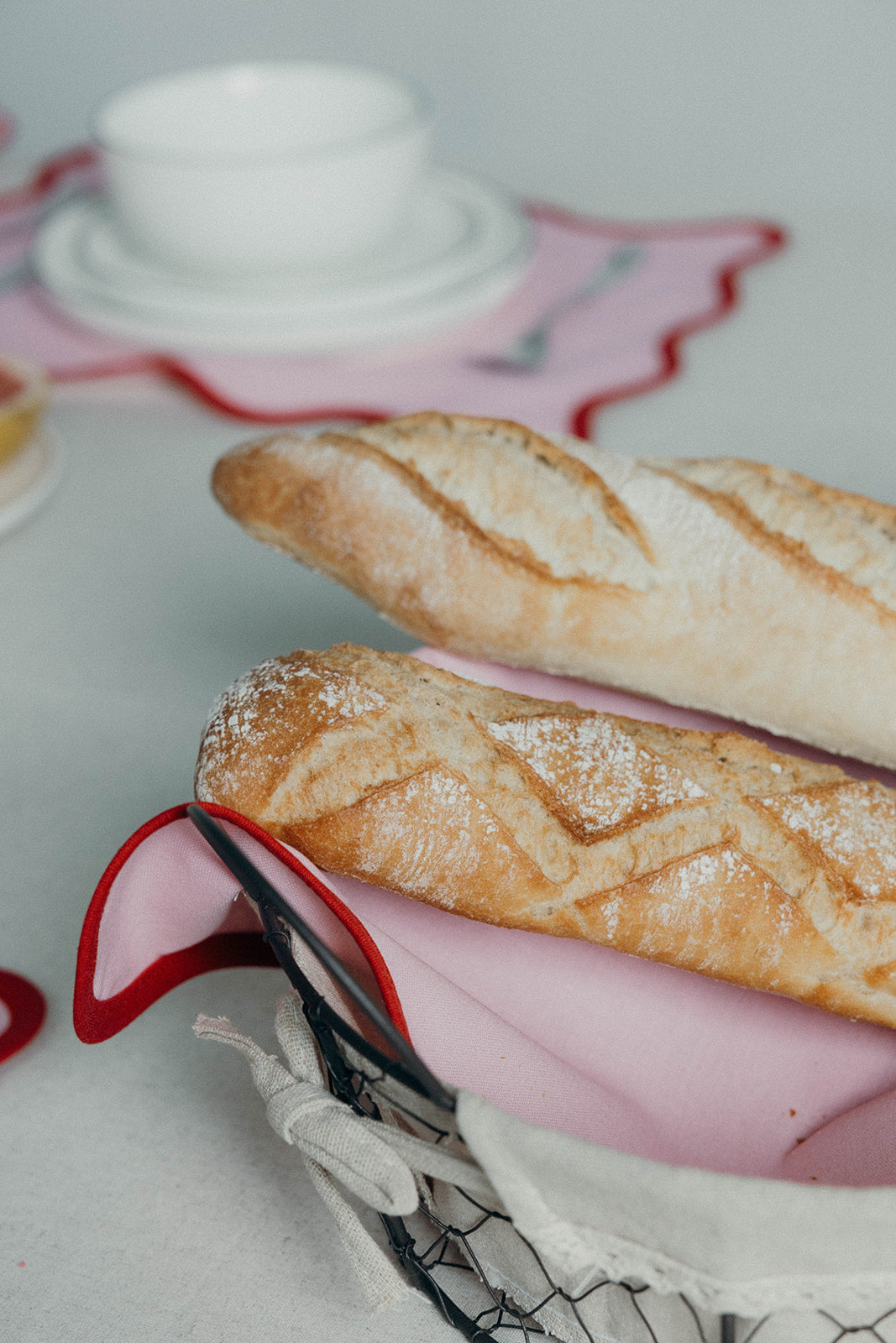 Fresh baguettes rest in a wire basket lined with a pink Lilou Scallop napkin at a softly blurred table setting.