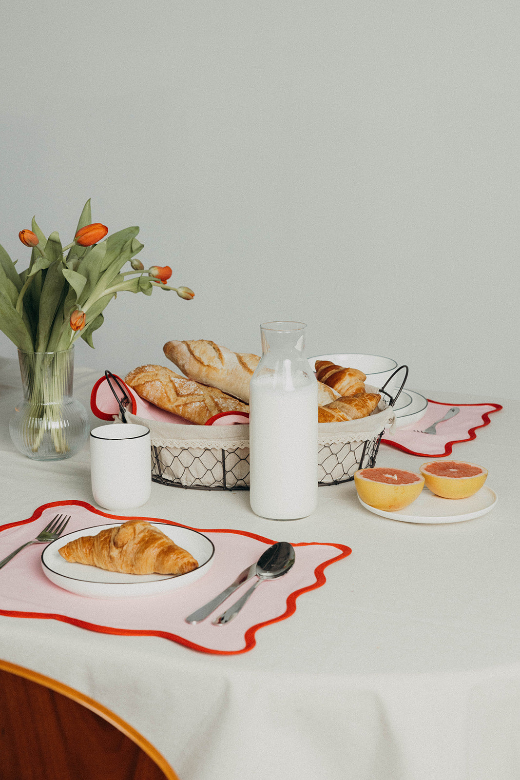 Pink Lilou Scallop placemat holds a plate with a croissant and cutlery beside a milk bottle and fresh tulips at a breakfast table.