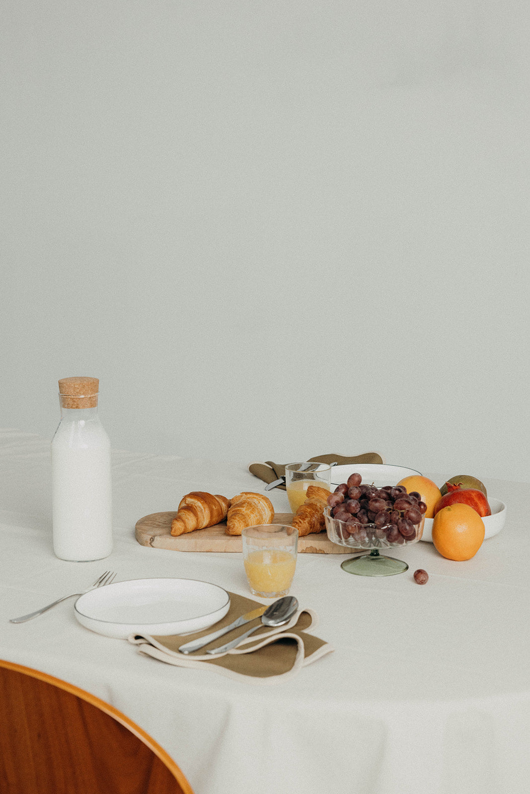 Olive Lilou Scallop placemat anchors a breakfast spread of croissants, grapes and orange juice on a neutral wood-backed table.