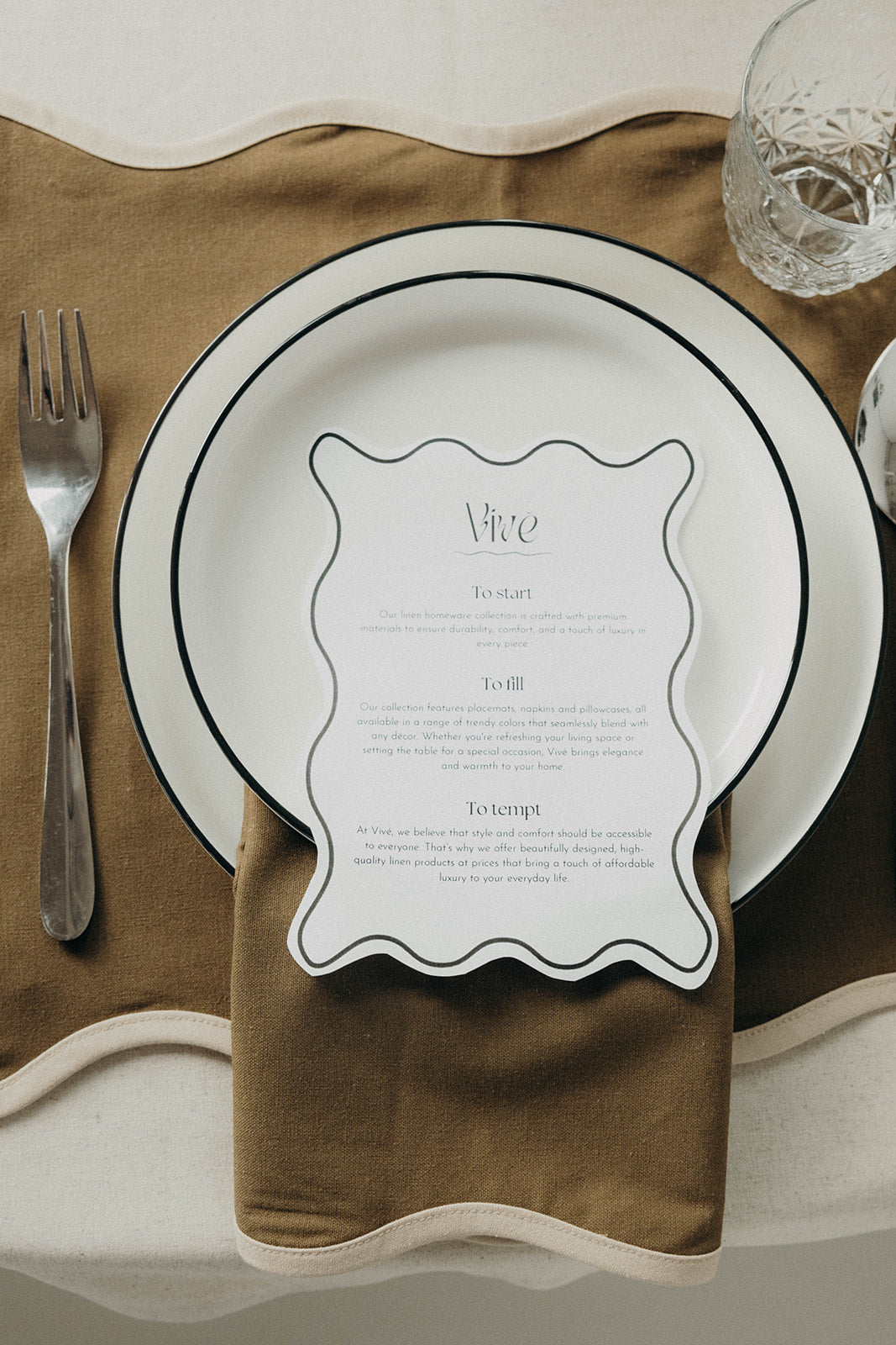 Close-up table setting showing the olive Lilou Scallop placemat, folded napkin and white plates with a Vivé menu card.