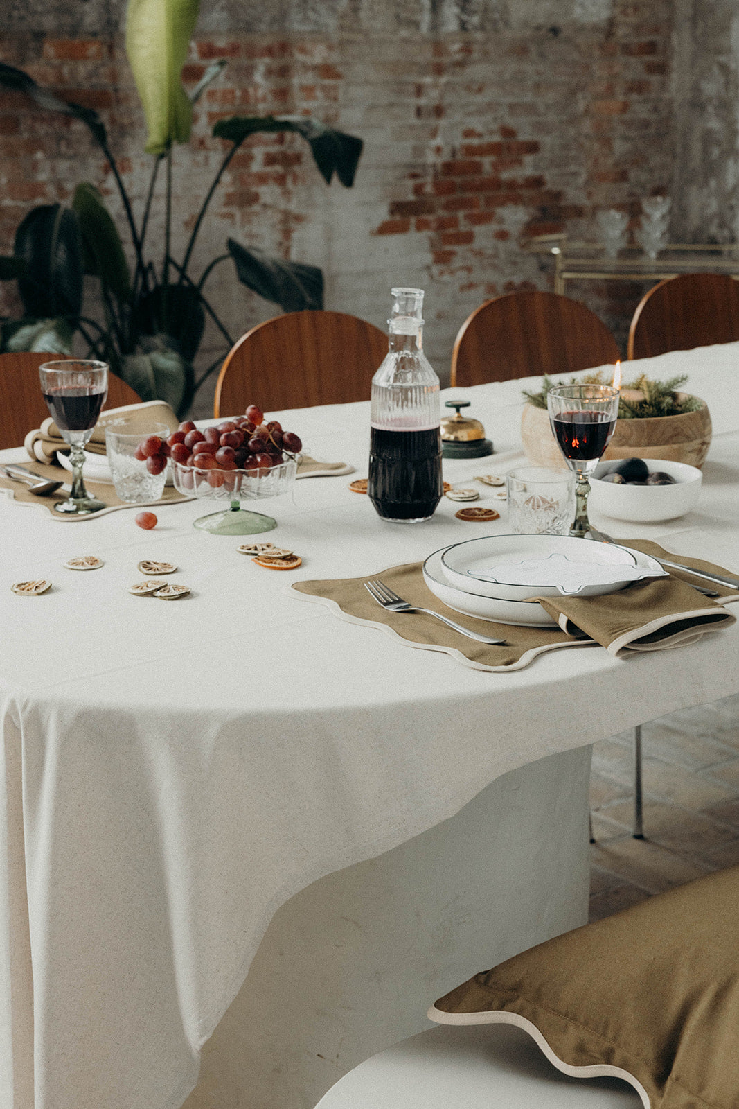 Olive Lilou Scallop placemat sets a long dining table with wine glasses, a lit candle and greenery against an exposed brick wall.