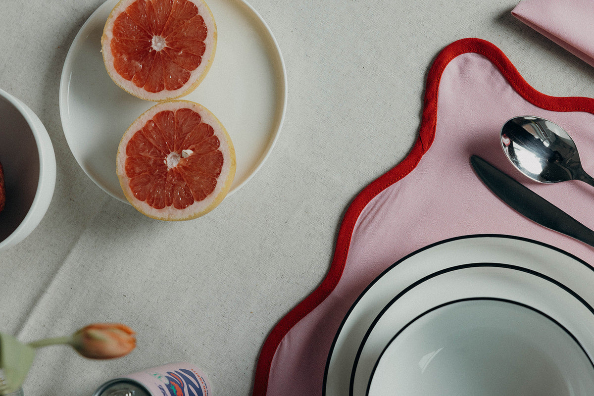 Orange Lilou Scallop placemat holds a plate of halved grapefruit beside lemons and a citrus cocktail on a white tablecloth.