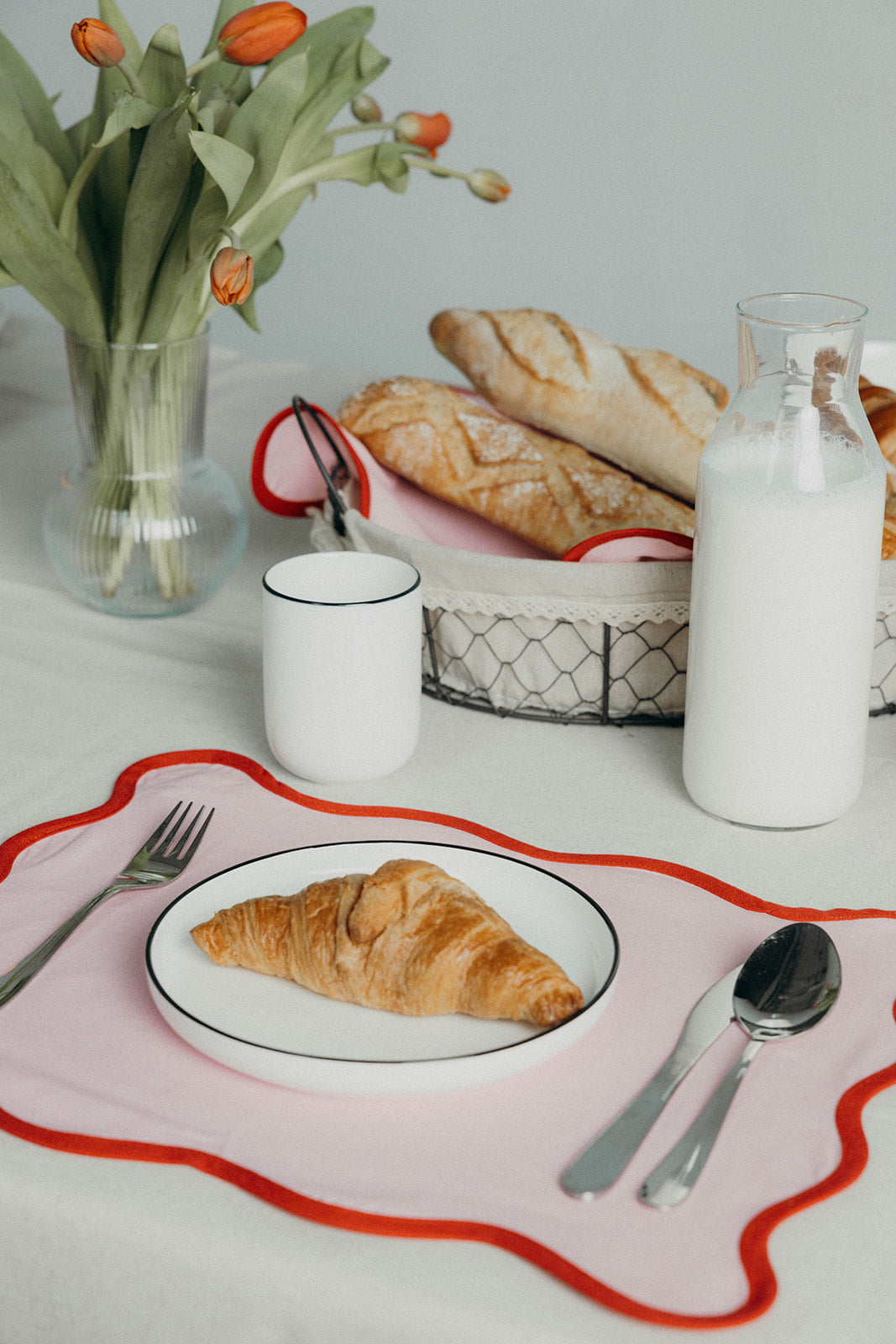 Pink Lilou Scallop placemat with a croissant on a plate sits beside a milk bottle, mug and basket of fresh baguettes.