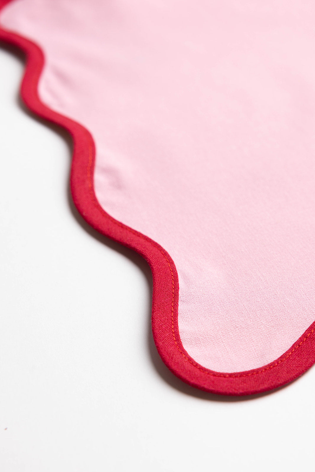 Close-up detail of the pink Lilou Scallop placemat's wavy red-trimmed border showing the cotton weave and stitching.