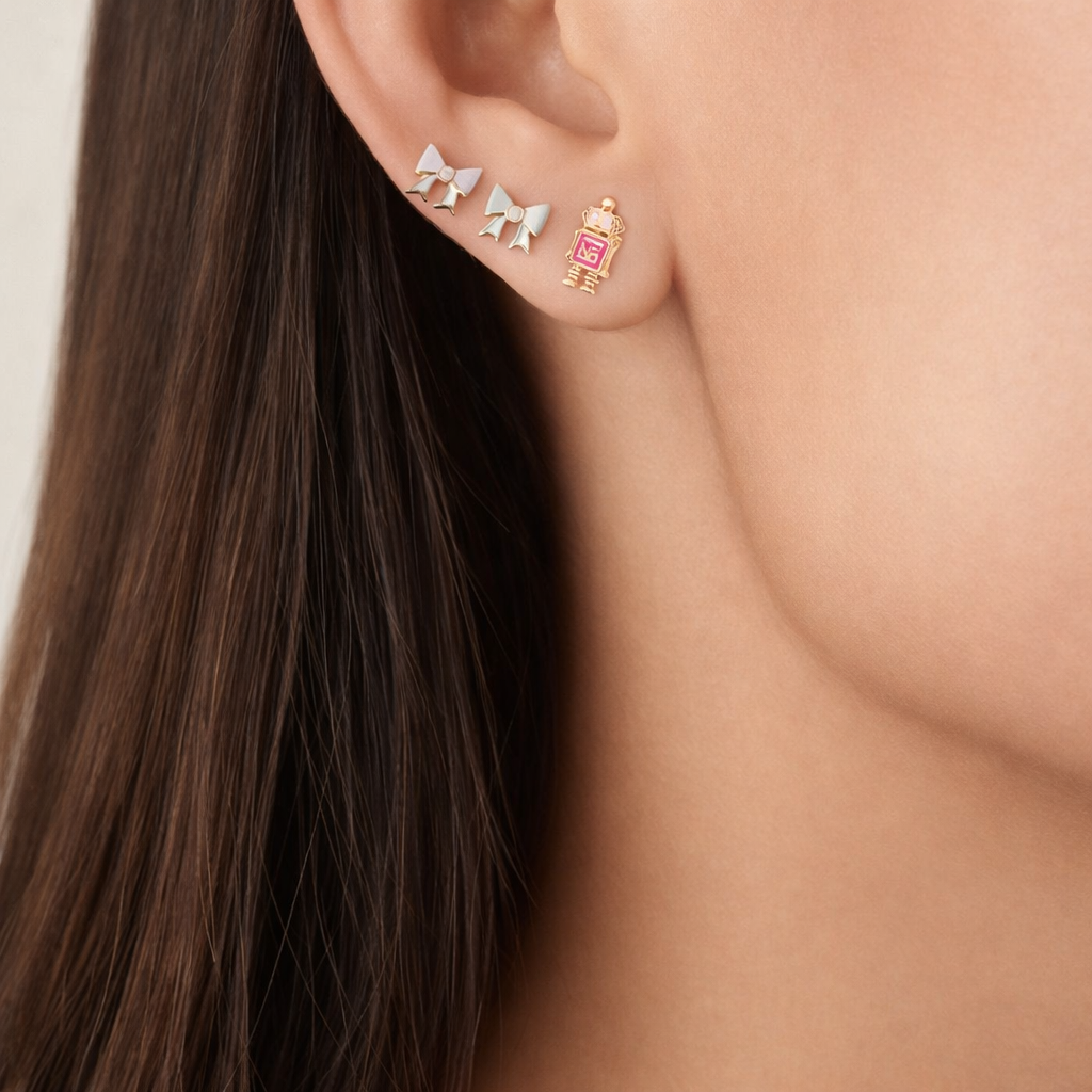 Worn layered with bow studs, Little Robot Earrings show their pink enamel version resting near the earlobe.