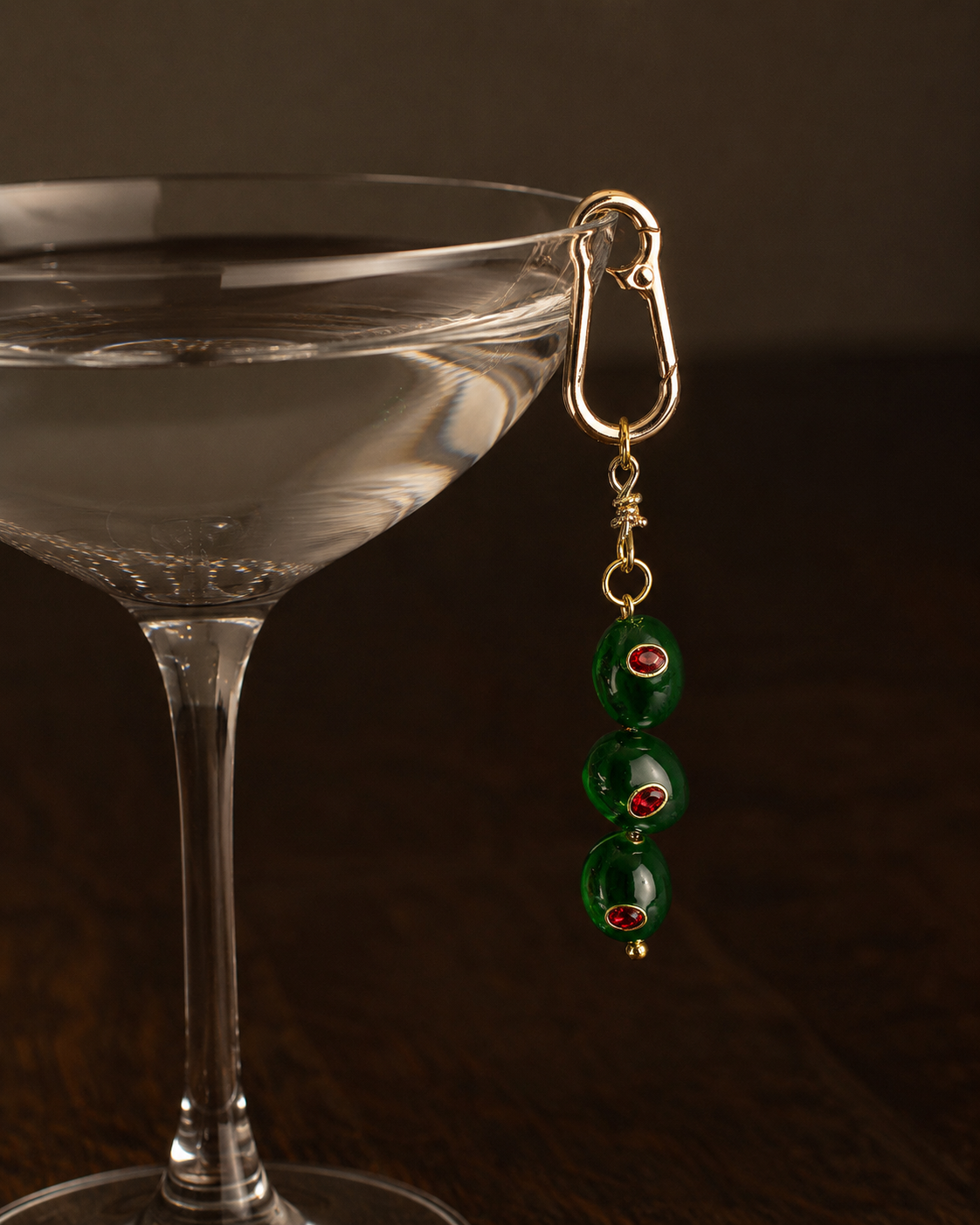 Folie à Trois Livia BAG CHARM, a gold clasp charm with three green beaded strands accented in red, hanging beside a cocktail glass.