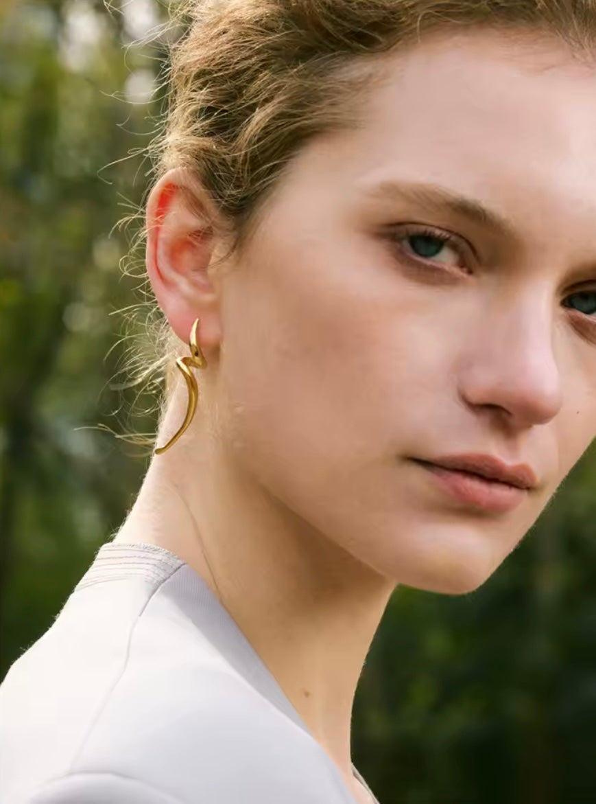 Lo Earrings worn on the ear, the gold curved swirl design catching light against loosely tied hair outdoors.