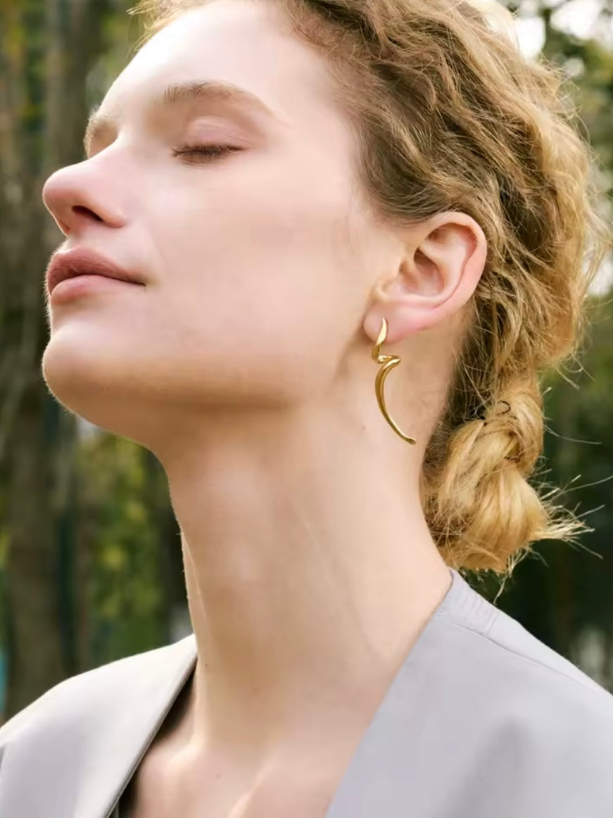 Lo Earrings worn in profile, the sculptural gold curl following the shape of the ear against a soft outdoor backdrop.