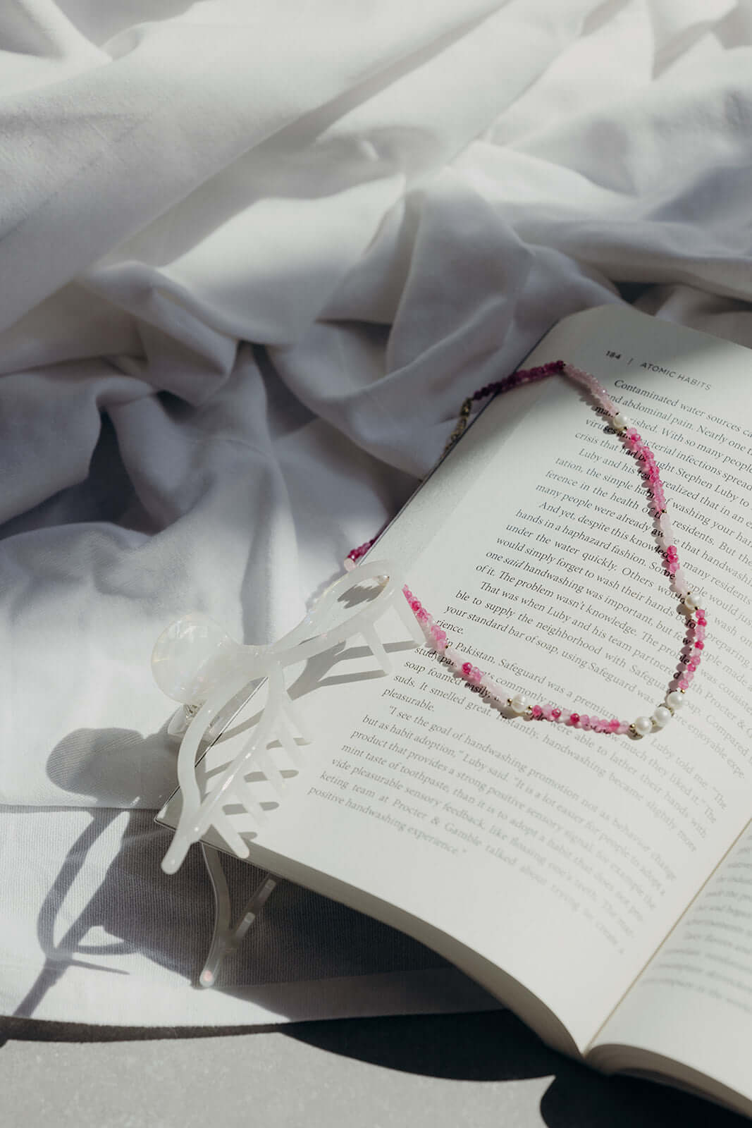 The Lolita NECKLACE styled on an open book, the pink and burgundy beaded strand draped over a jewelry display stand.
