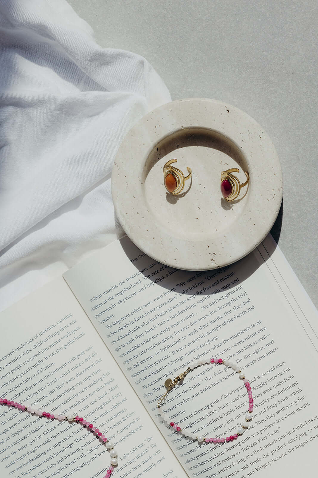 The Lolita NECKLACE styled on an open book beside a dish of gold gemstone earrings, in a sunlit flat lay scene.