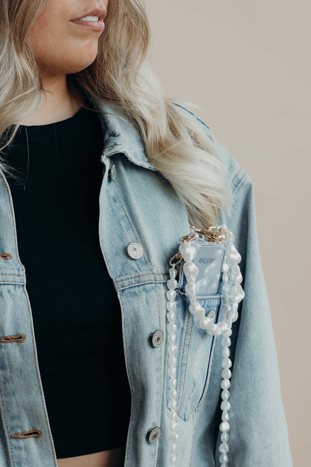 Phone with a long round pearl Long Pearl PHONE STRAP tucked into a denim jacket pocket, close-up on the model's face and hair.
