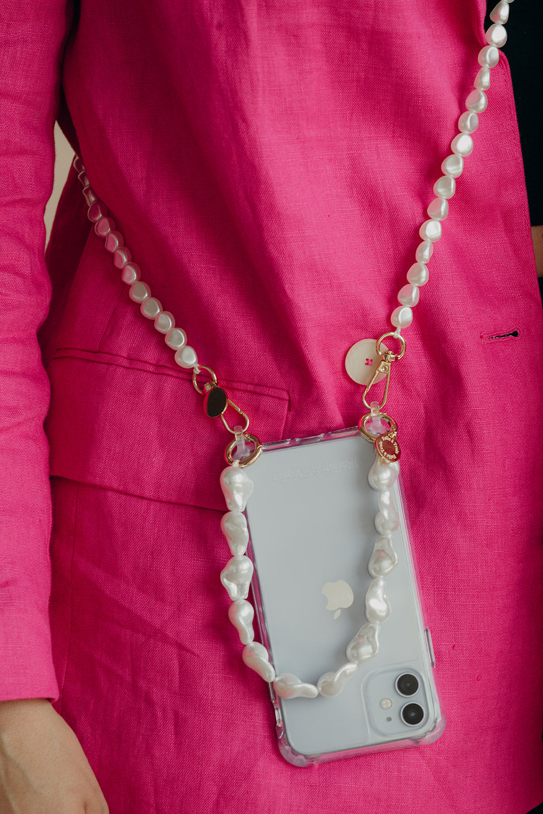 Phone in a clear case worn crossbody on a long white baroque pearl cord with a short pearl strap, against a bright pink linen suit.