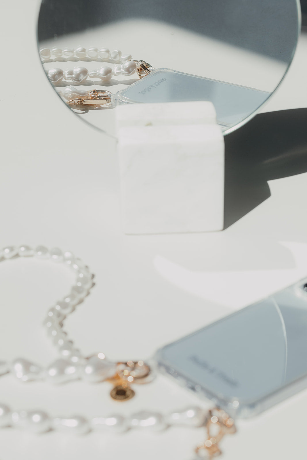 Long white pearl phone cord with gold clasps beside a clear phone case, a round mirror reflecting more pearls on a marble block.