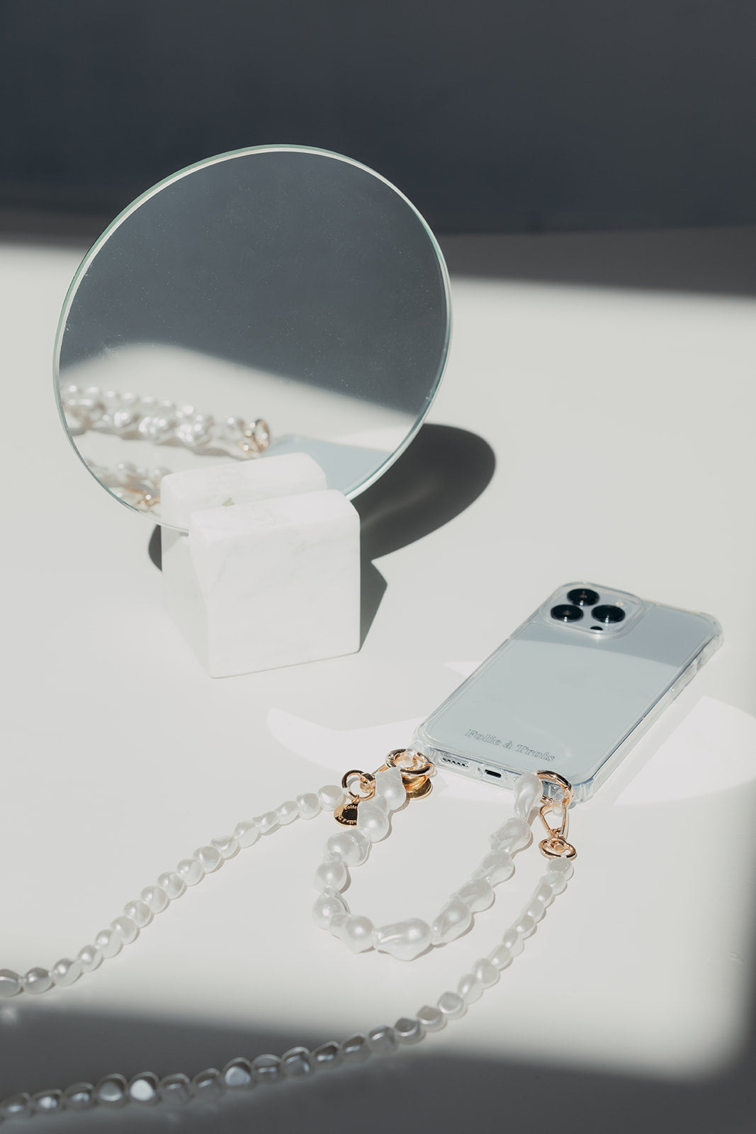 Clear phone case with a white pearl phone cord and gold clasps laid in sunlight beside a round mirror on a white surface.