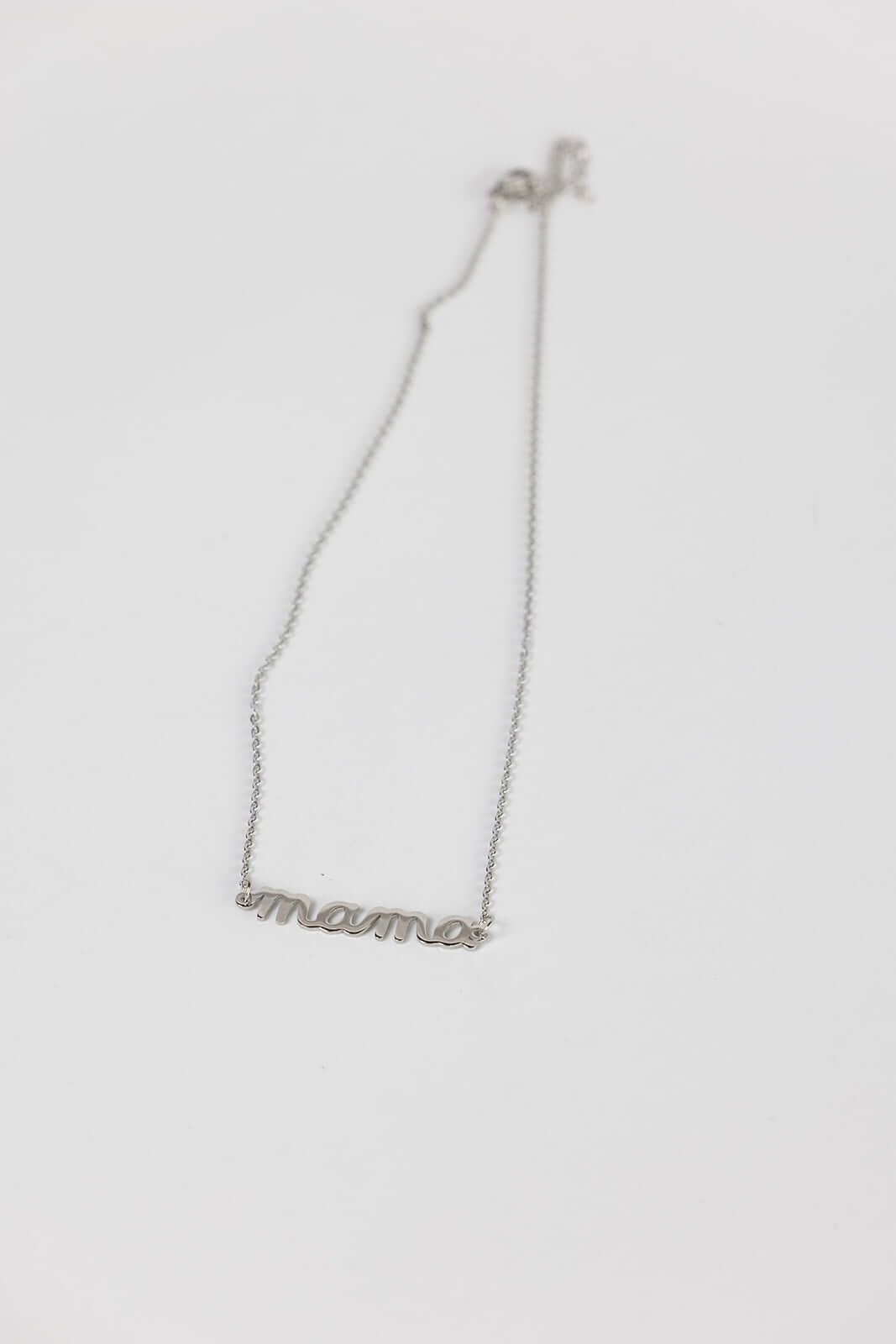 The Mama Big NECKLACE in silver, a script mama pendant on a fine chain, photographed lying on a white surface.