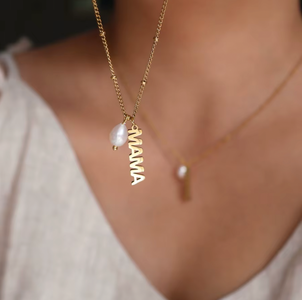 The Mama Pearl NECKLACE worn close-up at the neckline, its pearl drop and gold MAMA charm layered over a second delicate necklace.