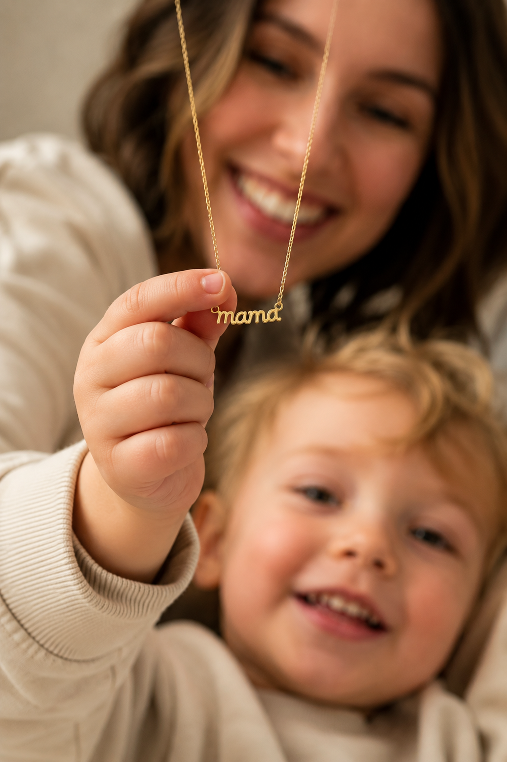 A smiling mother holds up the Folie à Trois Mama Small NECKLACE's script mama pendant while cradling her newborn wrapped in a knit blanket.