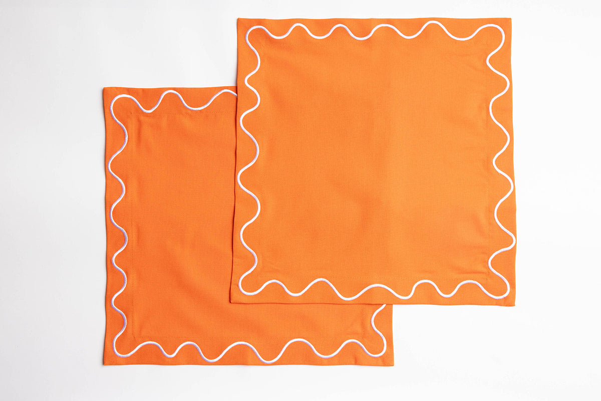 Two folded orange Maude napkins with white wave-trim edging, stacked flat on a white background.