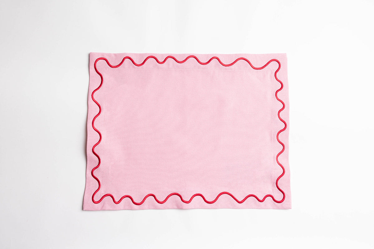 Pink Maude placemat with a red scalloped wave-trim border, laid flat on a white surface to show the embroidered edge detail.
