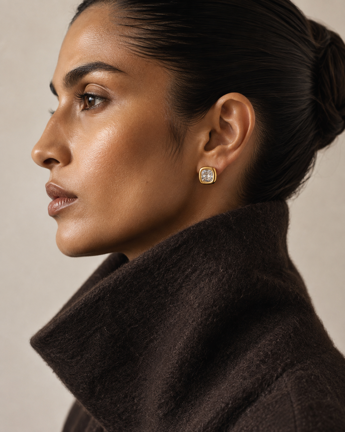 Profile view of a model in a brown coat wearing the gold Meridian EARRINGS, a cushion-cut zirconia bezel stud.