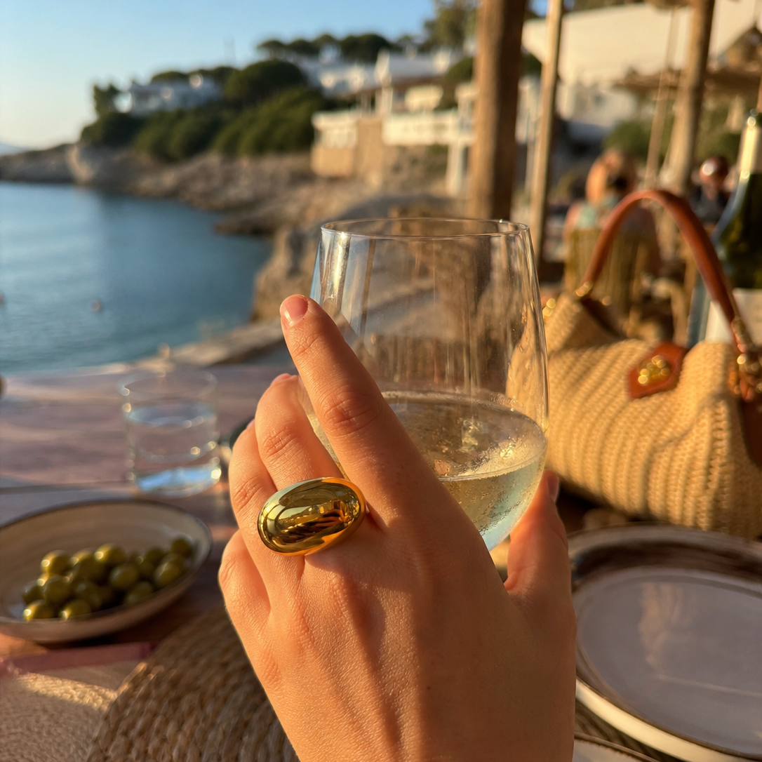 Model wears the gold Mirage RING while holding a wine glass at a seaside table with olives and a straw bag nearby.