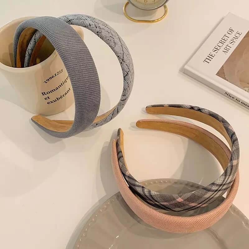 Two pairs of padded headbands - ribbed grey with grey tweed, and grey plaid with dusty pink - arranged near a mug and book.