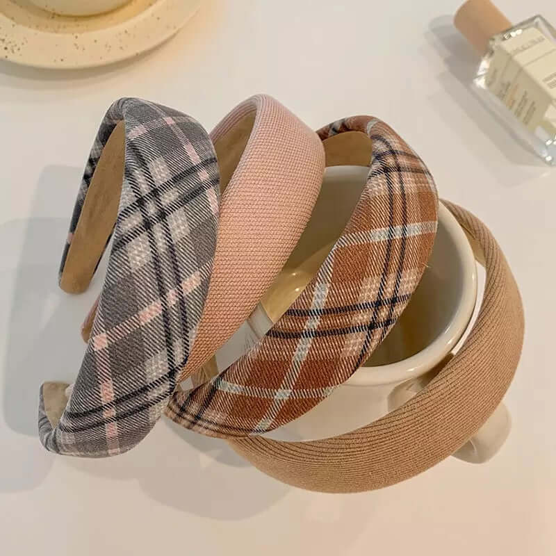 Four padded headbands in grey plaid, dusty pink, brown plaid and camel patterns stacked together over a mug.