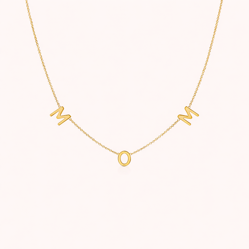 The Mom NECKLACE in gold, individual letters spelling MOM dangling from a fine chain, shown on a cream background.