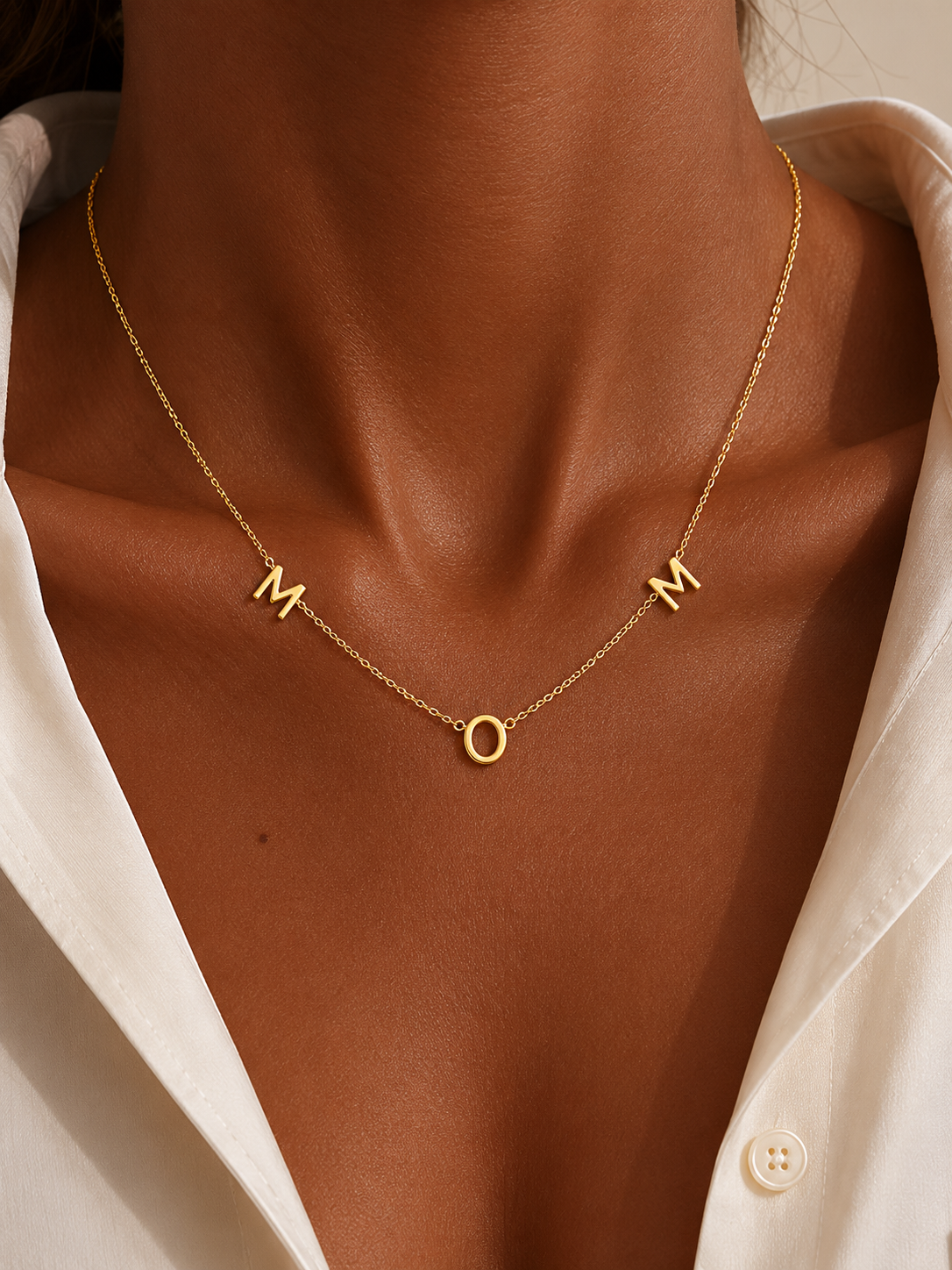 The Mom NECKLACE in gold worn at the neckline, individual gold letters spelling MOM above a white button-up shirt.