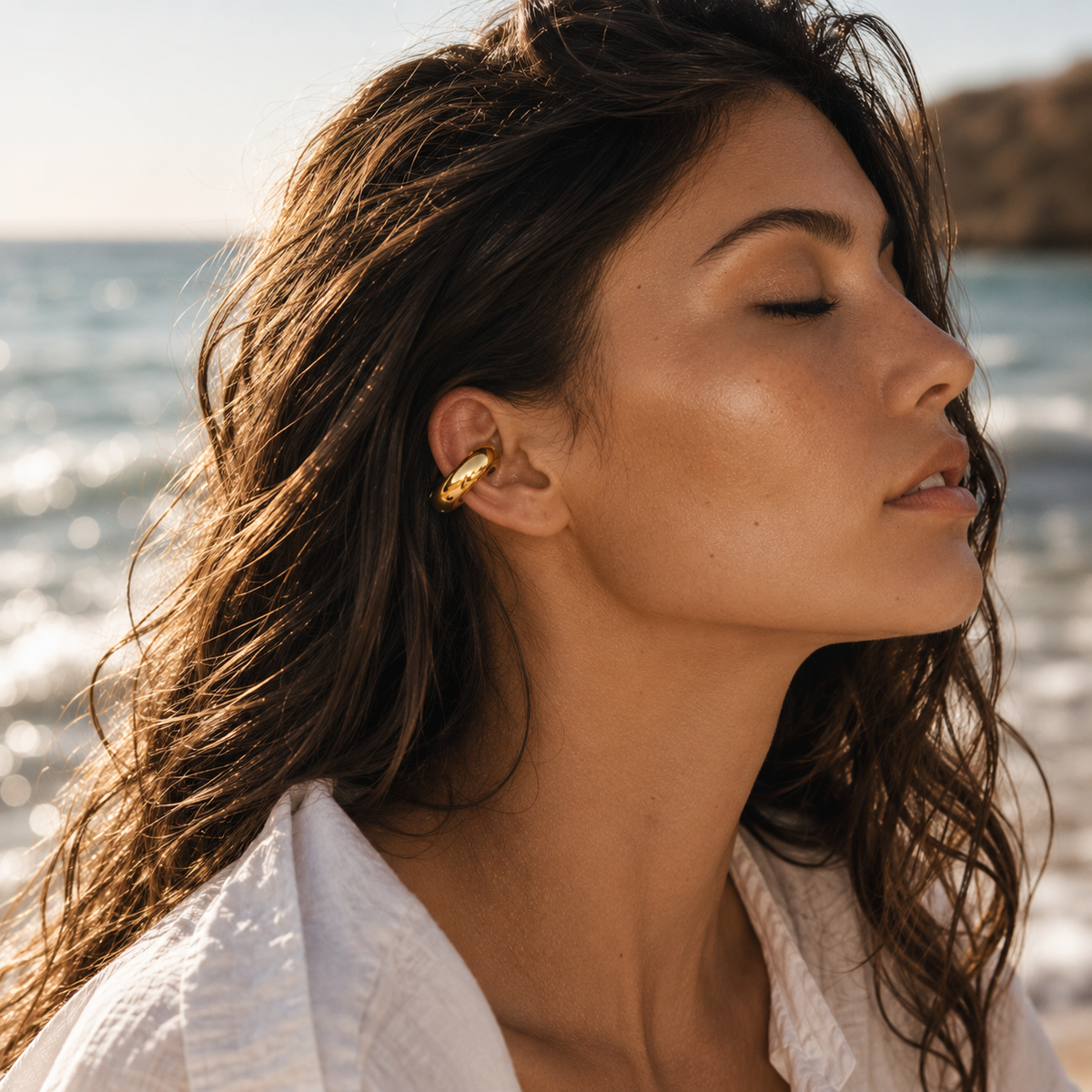 The Moon EAR CUFF worn on the ear at the beach, a smooth gold tube cuff on a model in a white shirt.