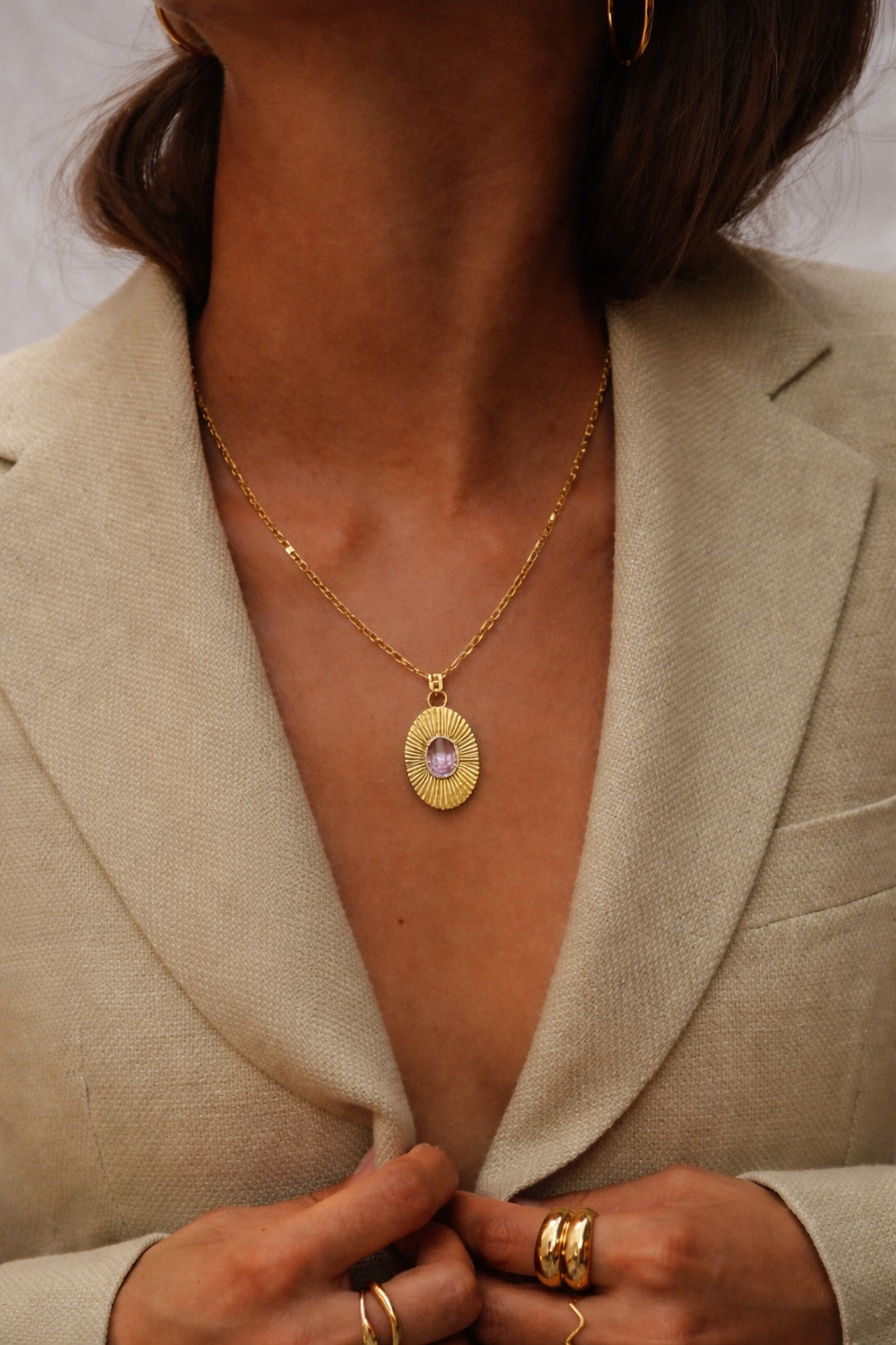 The More Than Friends NECKLACE in gold with a purple amethyst stone, worn at the neckline over a beige blazer.