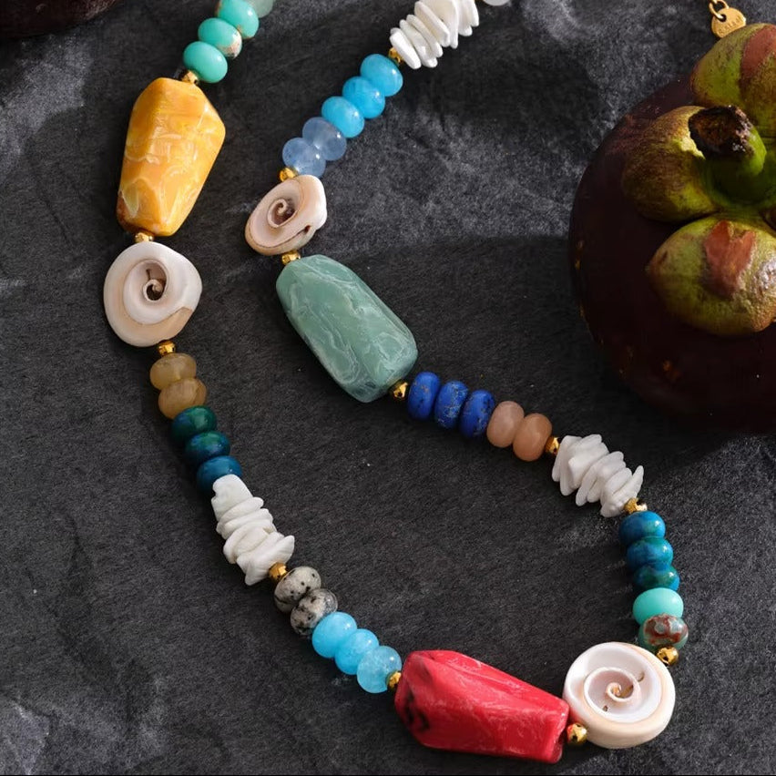 The Mykonos NECKLACE laid on dark stone beside a mangosteen fruit, its coral, amazonite, and carved shell beads in close detail.