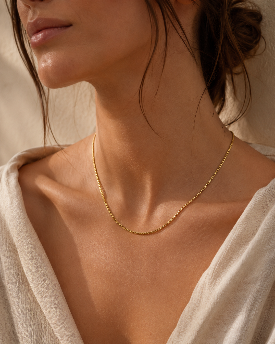 A model wears the gold Nara NECKLACE, a delicate box-chain necklace, layered close to the neckline over a linen top.