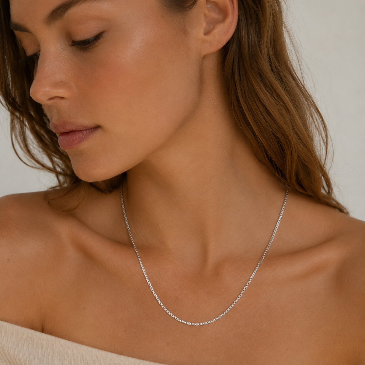 A model wears the silver Nara NECKLACE, a delicate box-chain necklace, layered close to the neckline over a cream top.