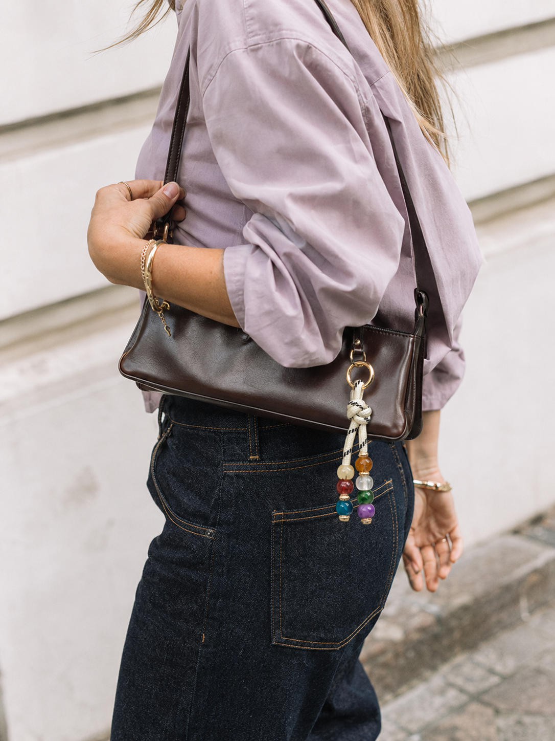 Folie à Trois Nodo BAG CHARM, a multicolor beaded strand on cream cord, clipped to a brown leather shoulder bag worn over denim.