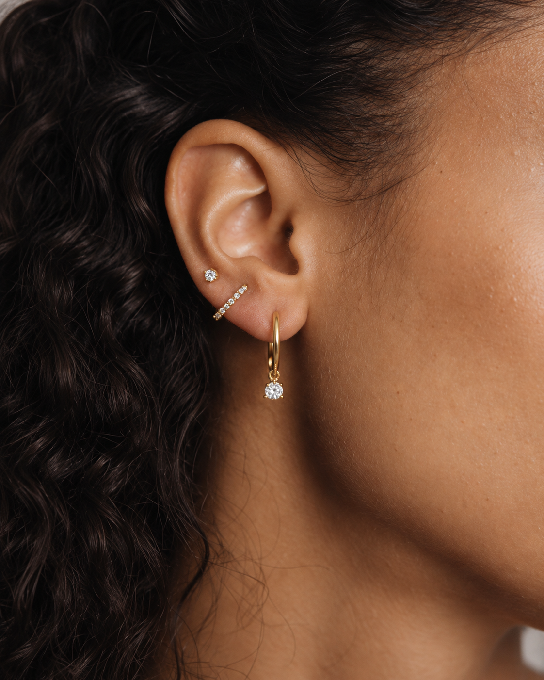 Model's ear wearing the gold Folie à Trois Noor SET stacked: a small stud, a pavé bar stud and a hoop with a dangling crystal.