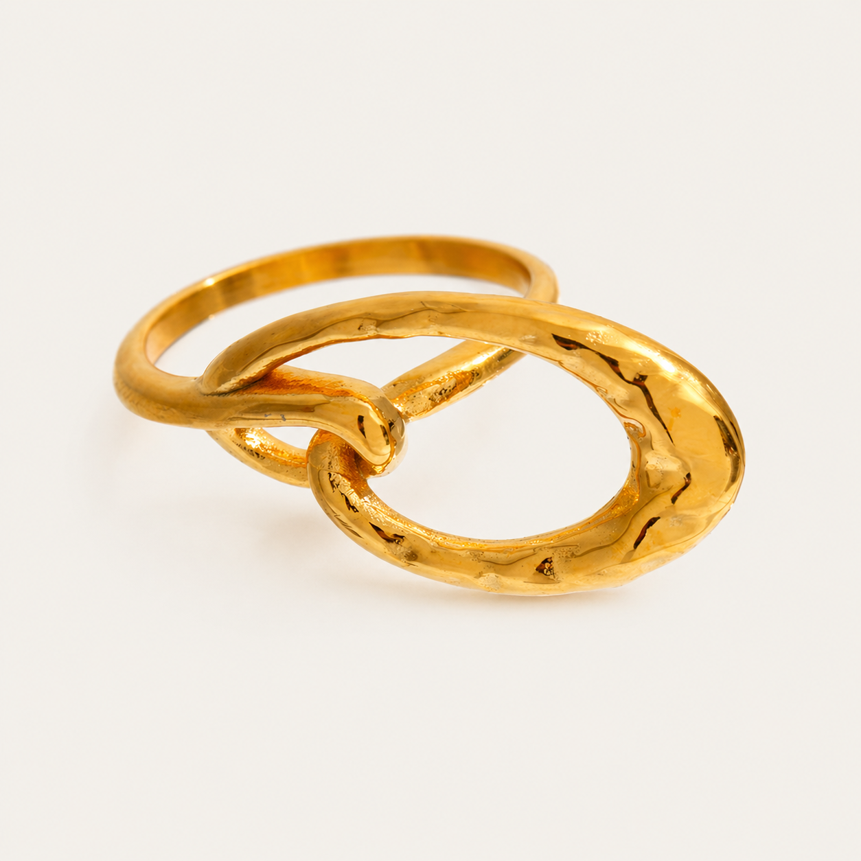 Gold ring where a smooth thin band interlocks with a hammered-texture open oval loop, shown on a cream backdrop — Folie à Trois Nova RING.