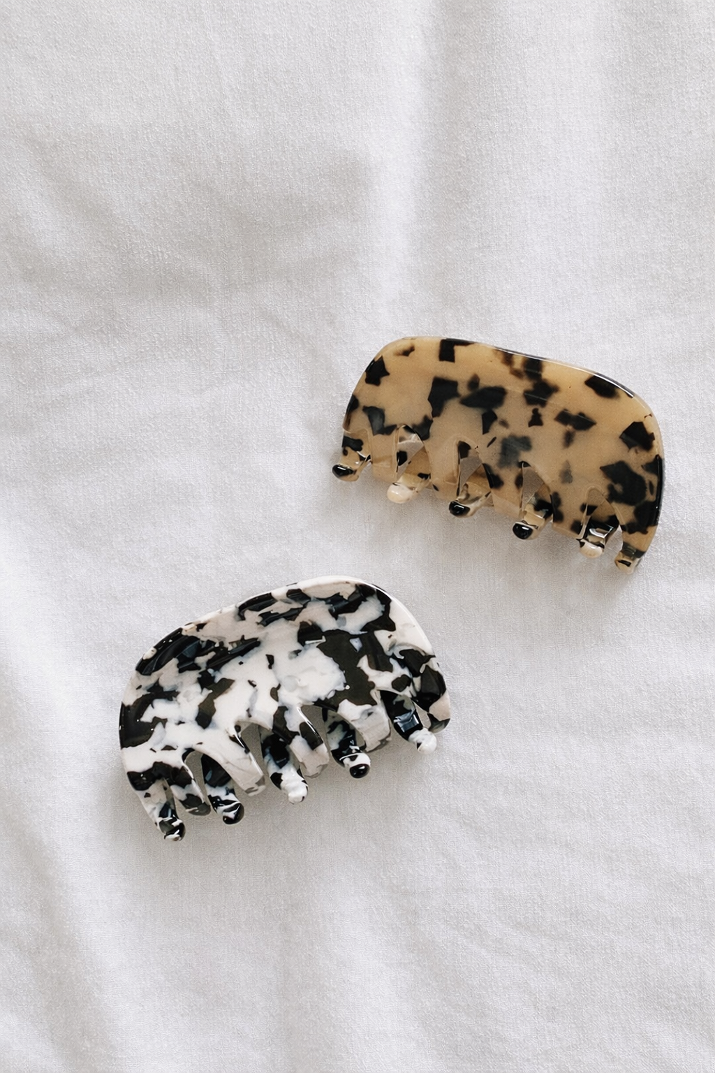 Two acetate hair claw clips, one tan tortoiseshell and one black and white mottled, resting together on white fabric.