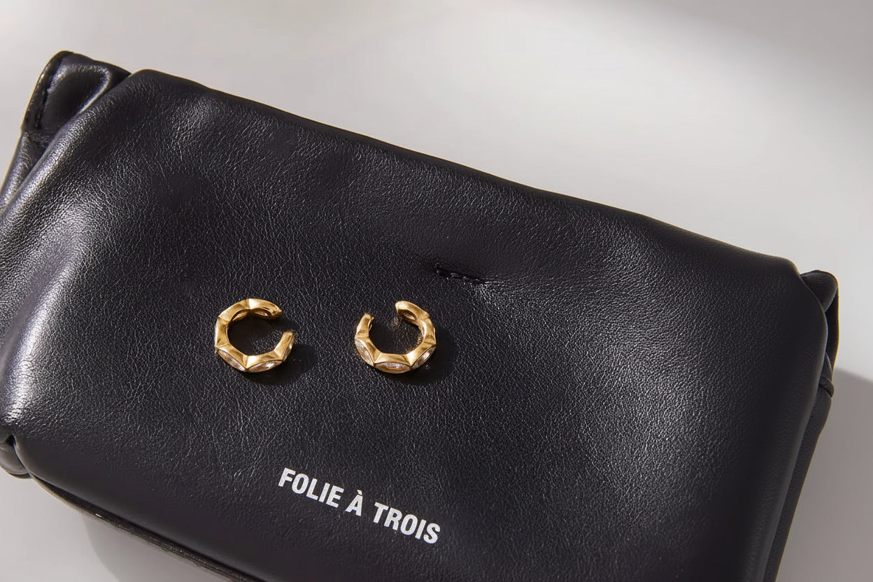 Pair of Overdrive ear cuffs resting open-face on a black pouch printed with the Folie a Trois logo, faceted gold bands.