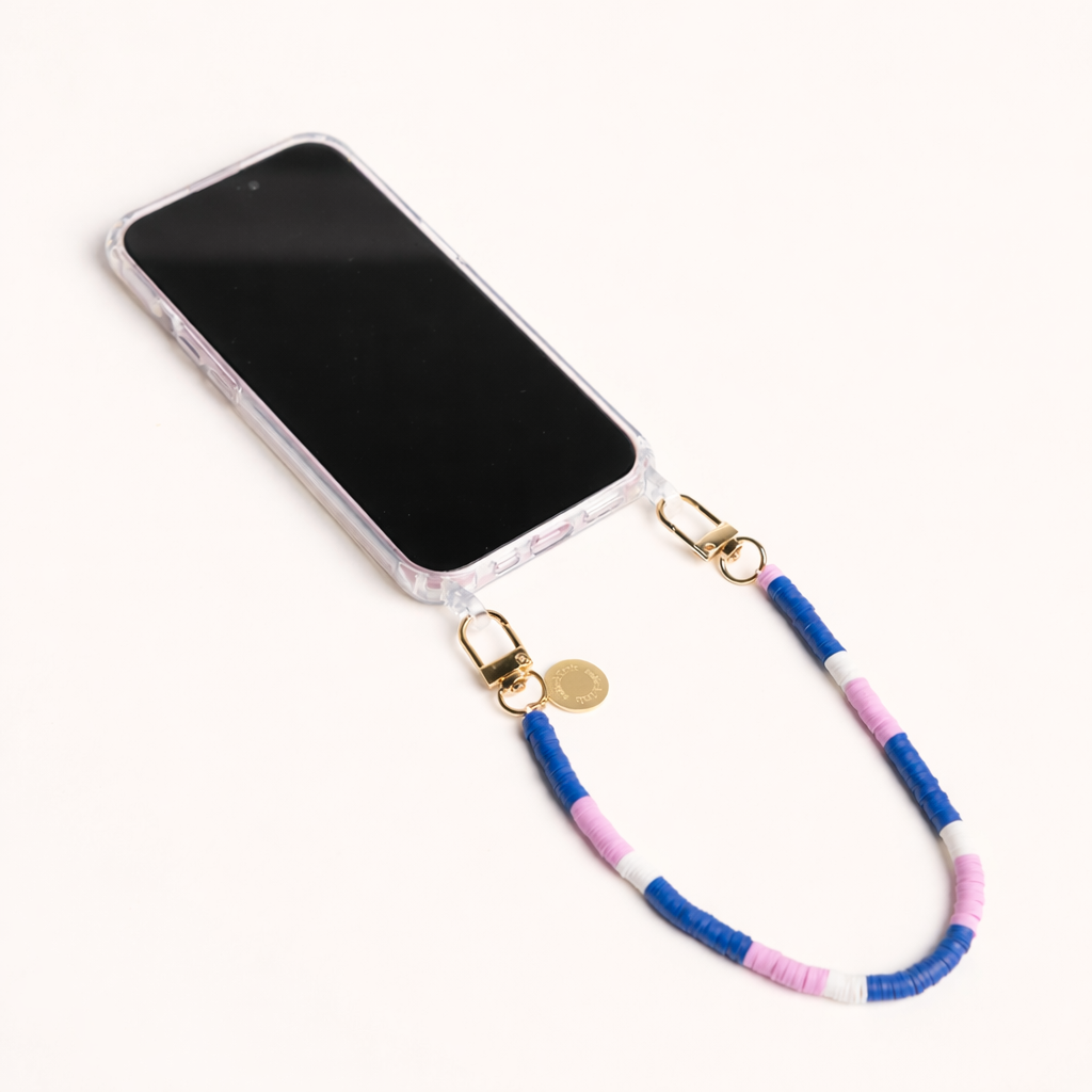 A clear phone case attached to a blue, pink, and white heishi bead phone cord with a gold clasp and engraved gold charm, shown on a cream background.
