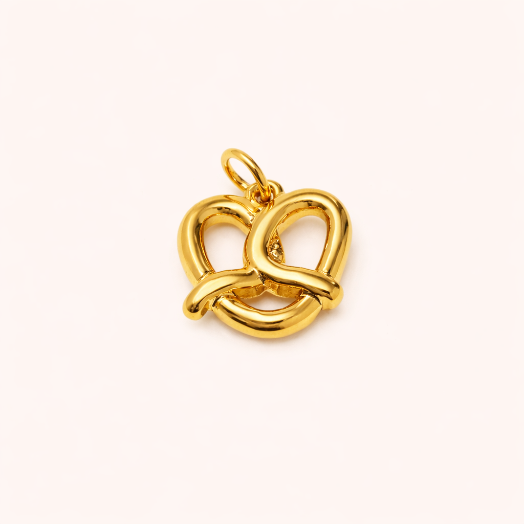 The Pretzel CHARM in a studio flat lay, a polished gold pendant shaped like a soft pretzel on a cream background.