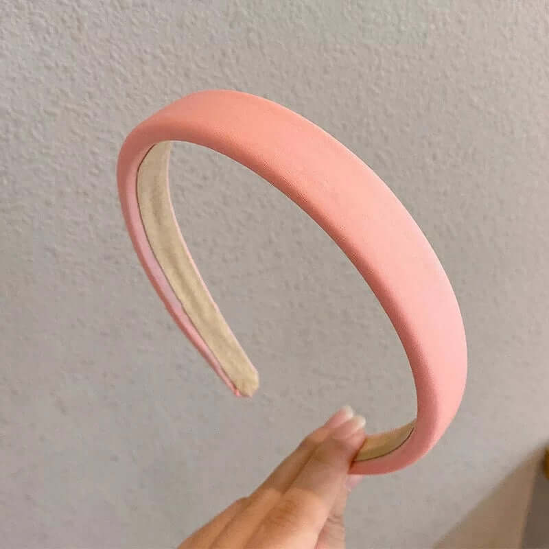 A single coral peach velvet-look padded headband held between fingers against a plain wall.